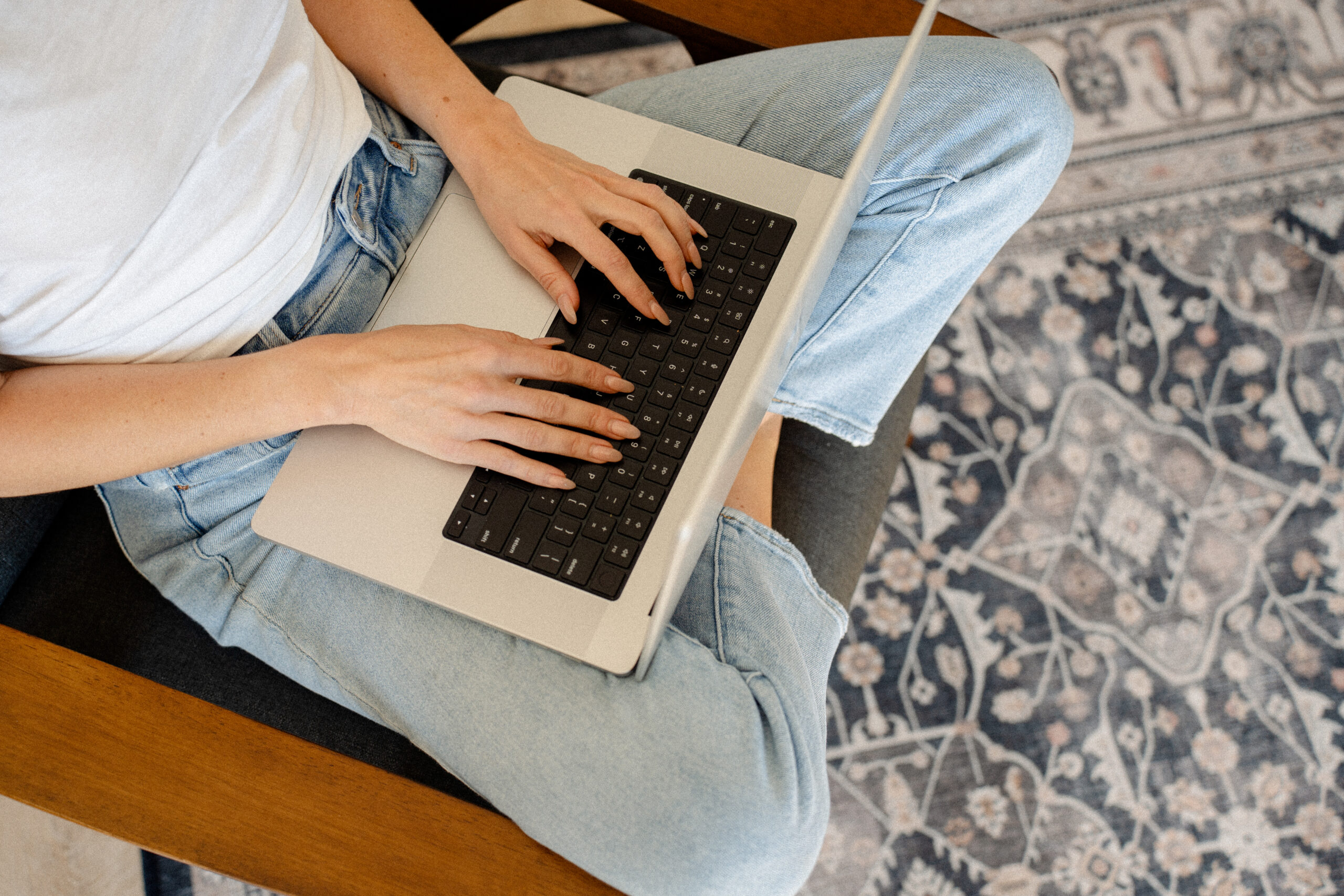 Woman sitting on a chair in bluejeans typing on a silver computer working on her creative business.