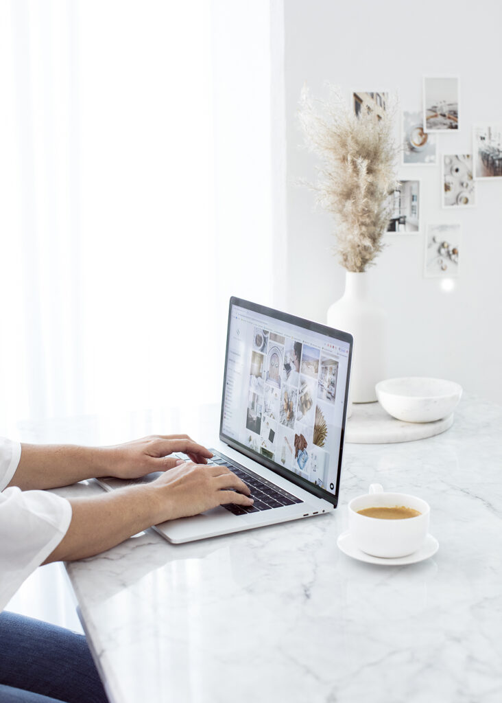 A woman types on a laptop in a white room on a marble counter top as she collects images to customize her Lily template for her new Showit Website.