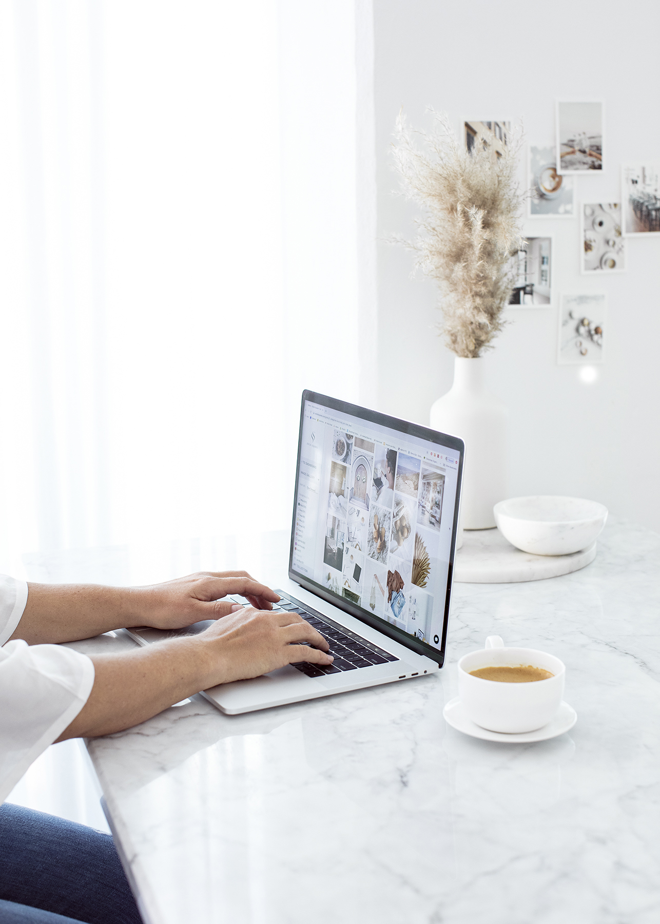 A woman types on a laptop in a white room on a marble counter top as she collects images to customize her Lily template for her new Showit Website.